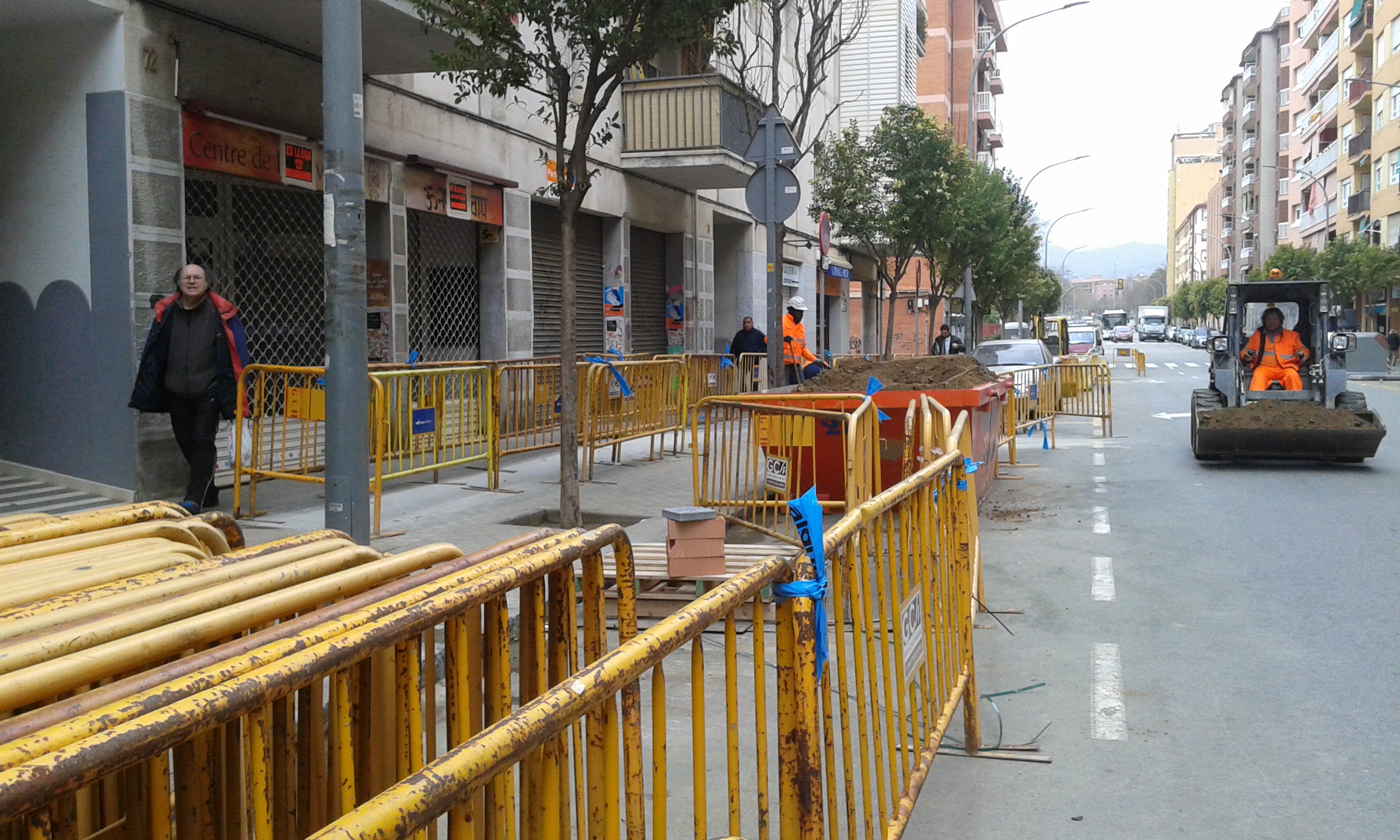 Carrer Biada amb tanques d'obra a l'esquerra, operari treballant amb excavadora a la calçada i vianants caminant per la vorera