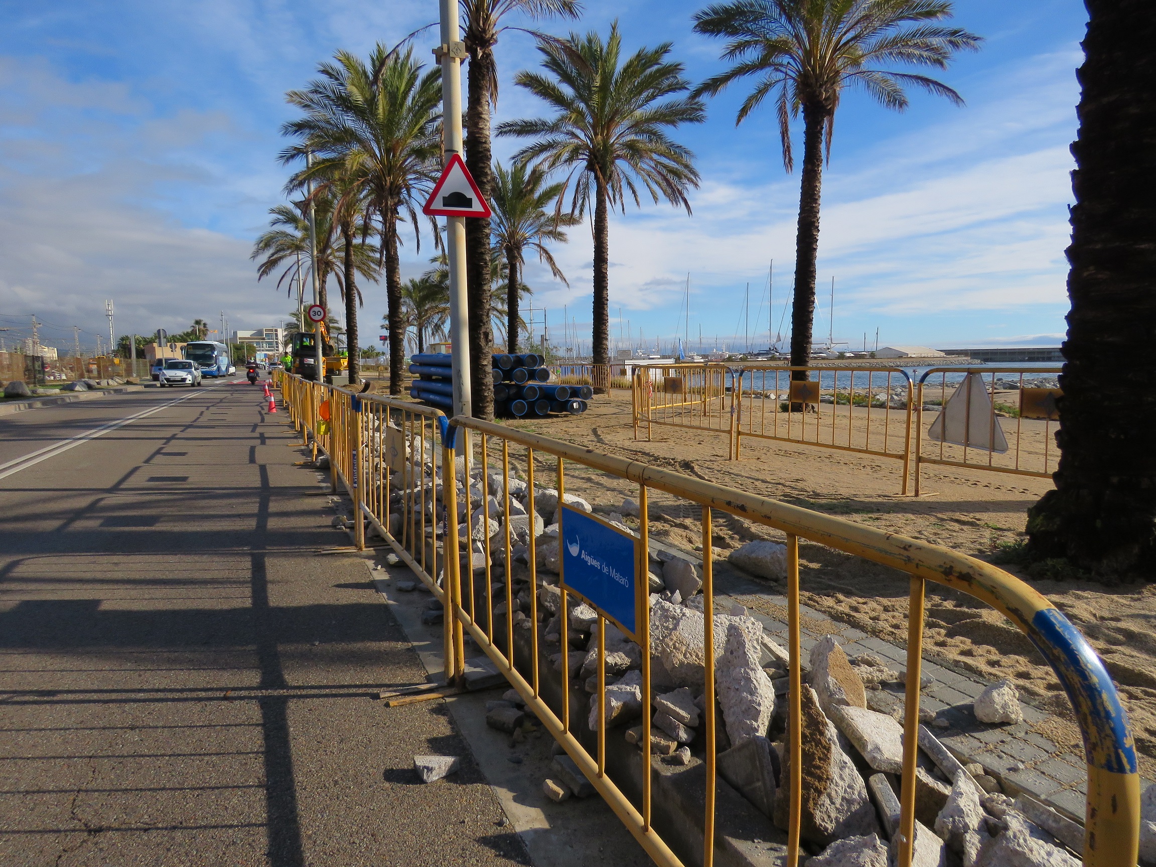 Avinguda del Port amb rasa i pedres, oberta a la dreta, protegida per tanques. Palmeres i platja al fons