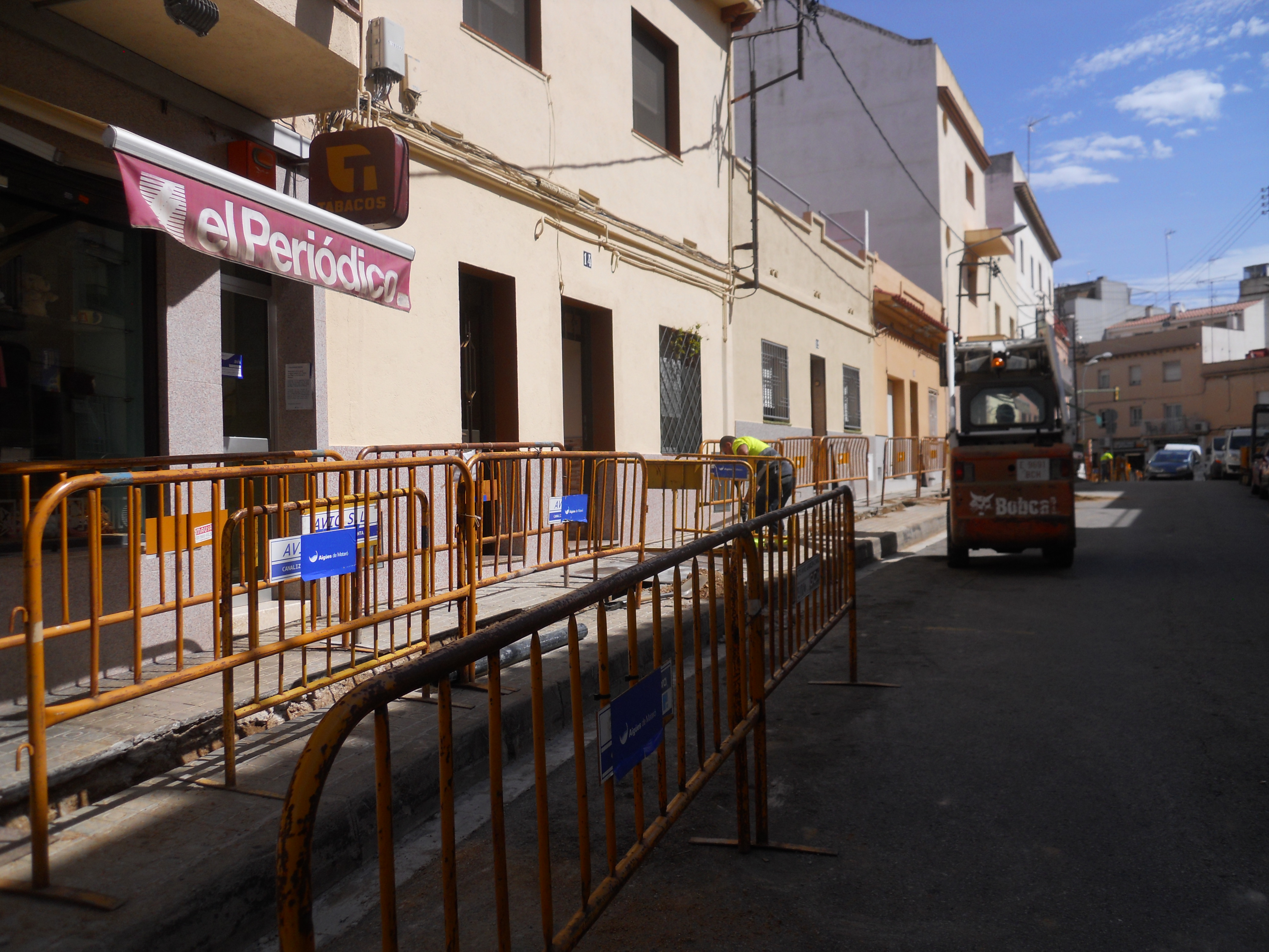 carrer en obres amb vorera protegida per tanques d'Aigües de Mataró, operari treballant en una rasa i excavadora circulant pel centre de la calçada