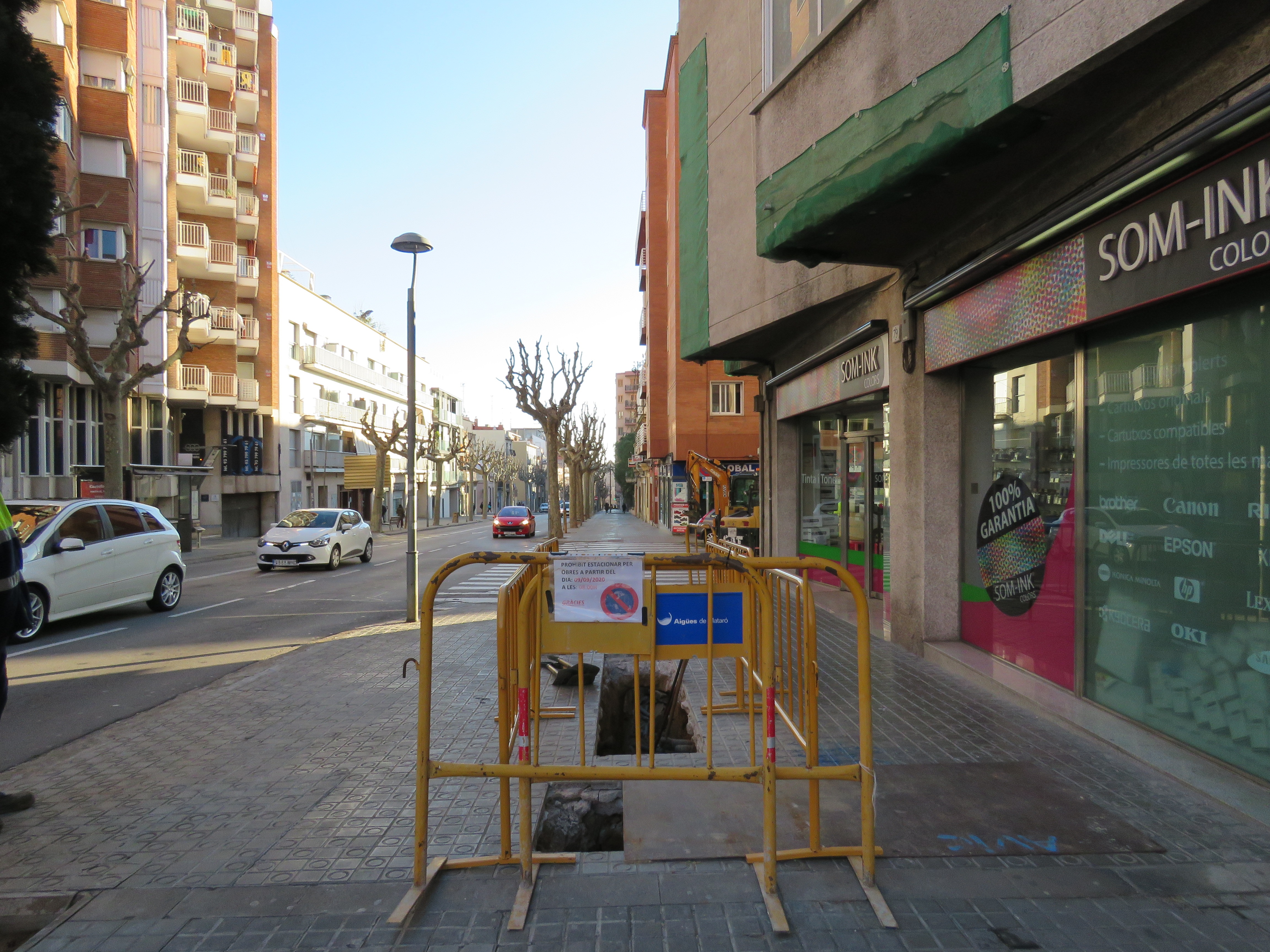 Carrer de Jaume Isern amb cotxes circulant per la calçada a l'esquerra i tanca d'obres protegunt una rasa oberta a la vorera