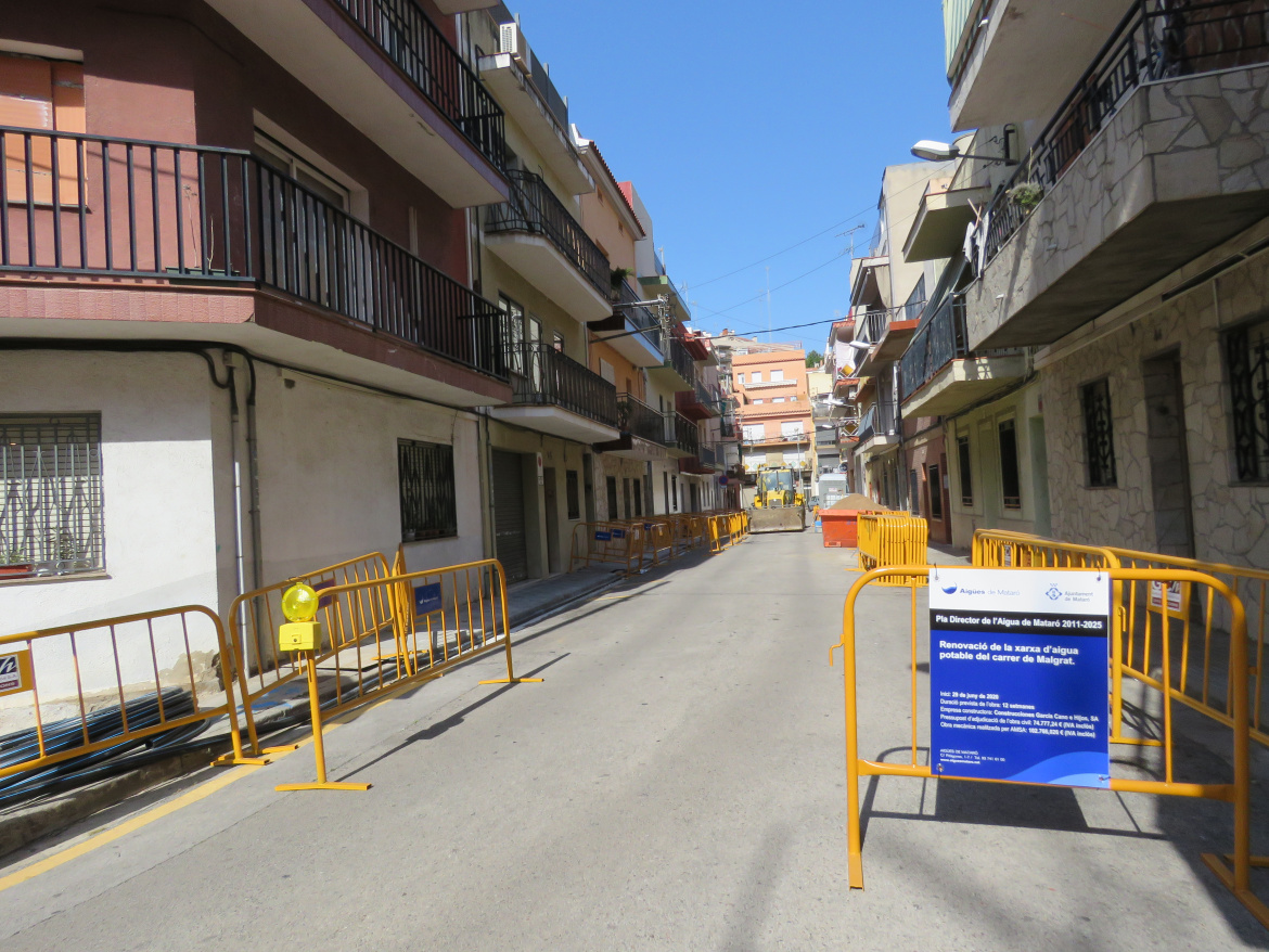 Carrer de Malgrat amb cartell d'inici d'obres en primer pla i operari treballant amb una excavadora al fons