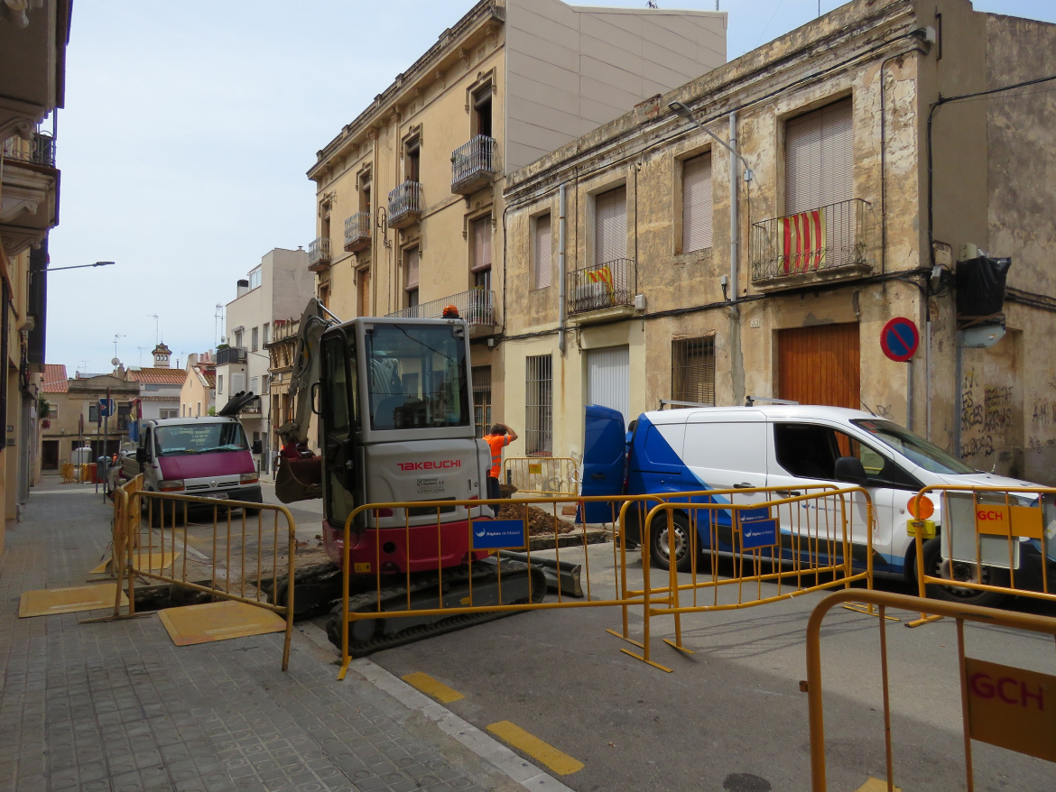 Màquina excavadora en primer pla amb furgoneta d'Aigües de Mataró aparcada a la vorera del costat