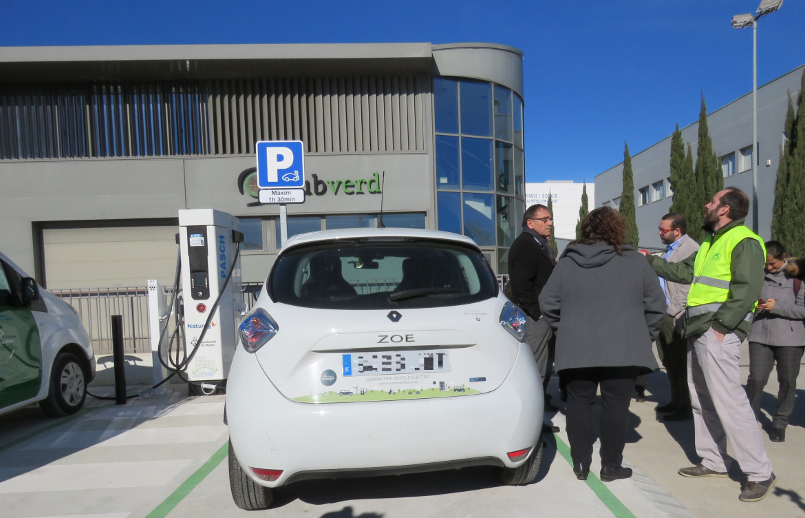 Cotxe elèctric davant de l'edifici de Tubverd connectat al punt de recàrrega amb persones al costat dret.jpg