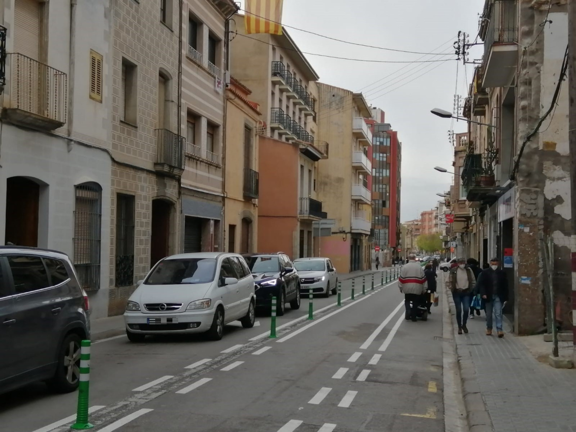 Camí Ral amb cotxes circulant per la calçada i vianants caminant per la vorera i tram de la calçada protegit per pilones verdes