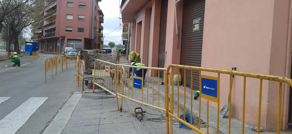Dos operaris d'Aigües de Mataró obrint una rasa amb una màquina, protegits per tanques d'obres
