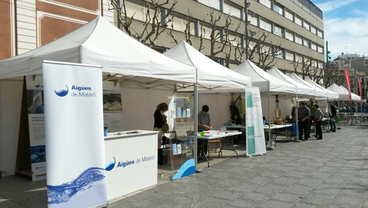 Expositors blancs a la plaça de Santa Anna amb persones a dins. en primer pla, expositor i cartell d'Aigües de Mataró.jpg